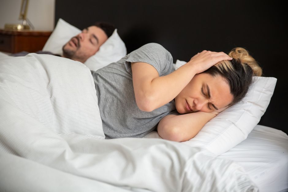 pexels-photo-7556638-7556638 A woman looks frustrated in bed as her partner snores loudly, highlighting sleep disturbance issues.